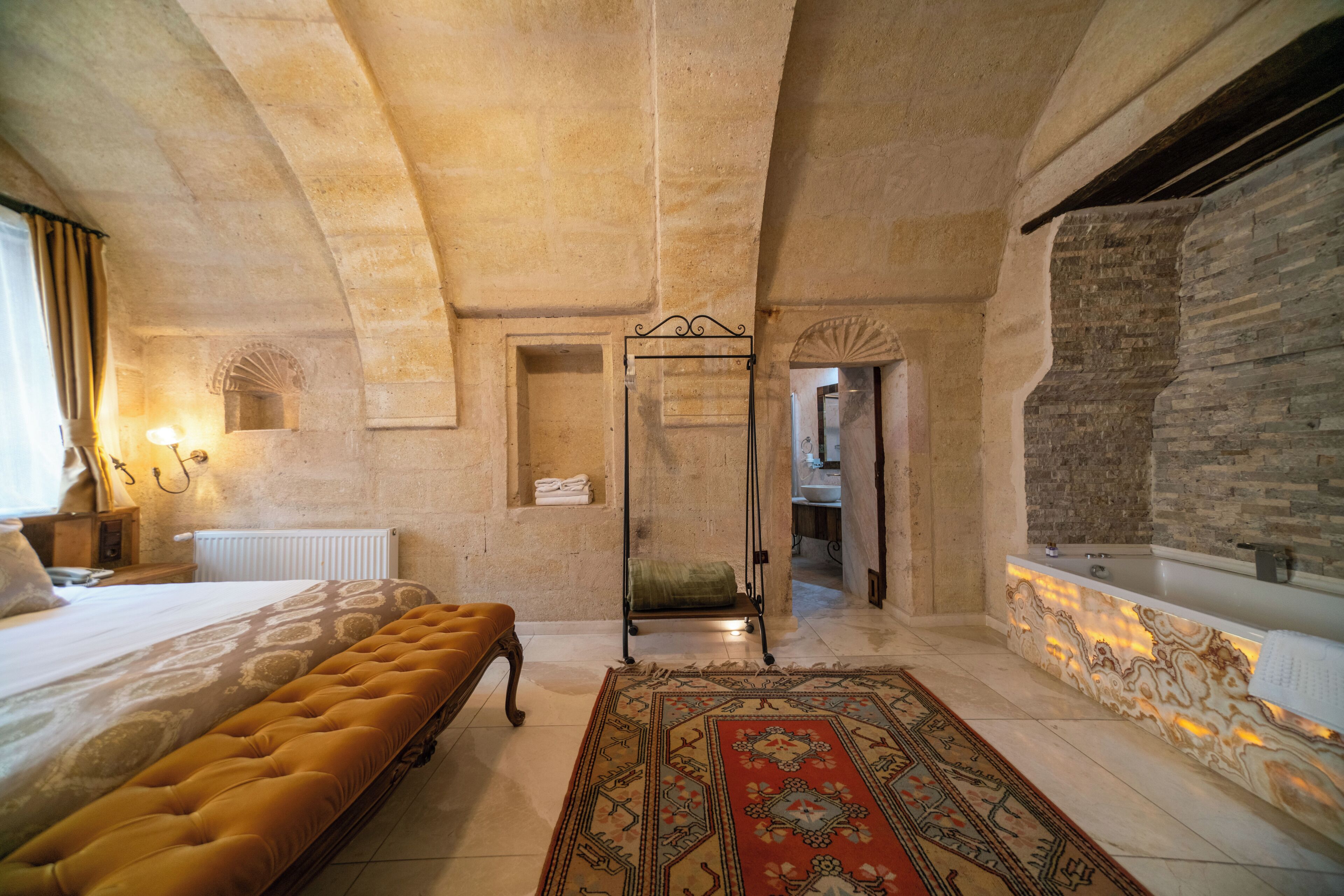 Une pièce comprenant un lit, un banc, un tapis à motifs, un mur en pierre et une salle de bain avec une baignoire en marbre.