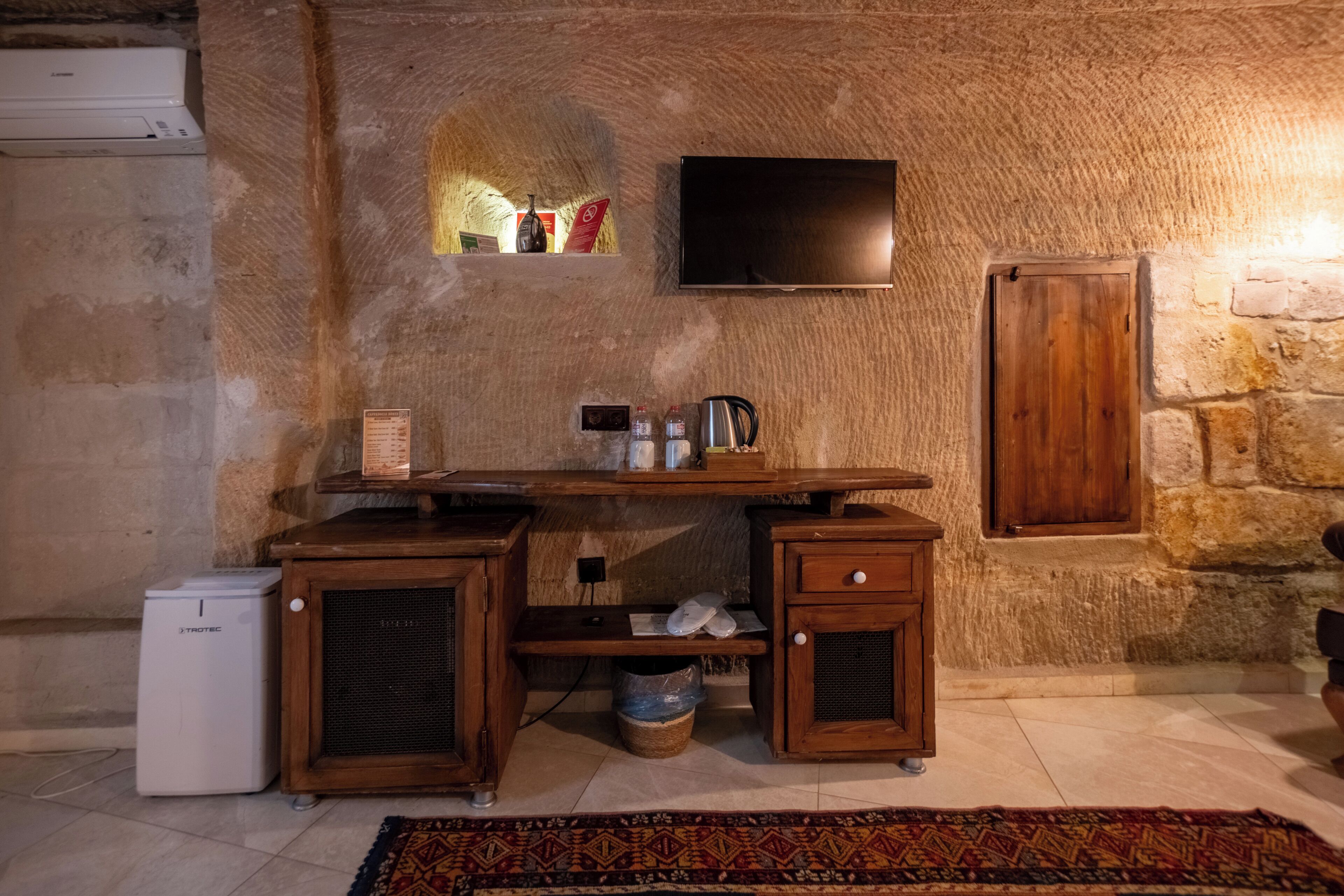 A rustic room with a stone wall, a flat-screen TV mounted above a wooden desk, a small refrigerator, and a wooden cabinet.