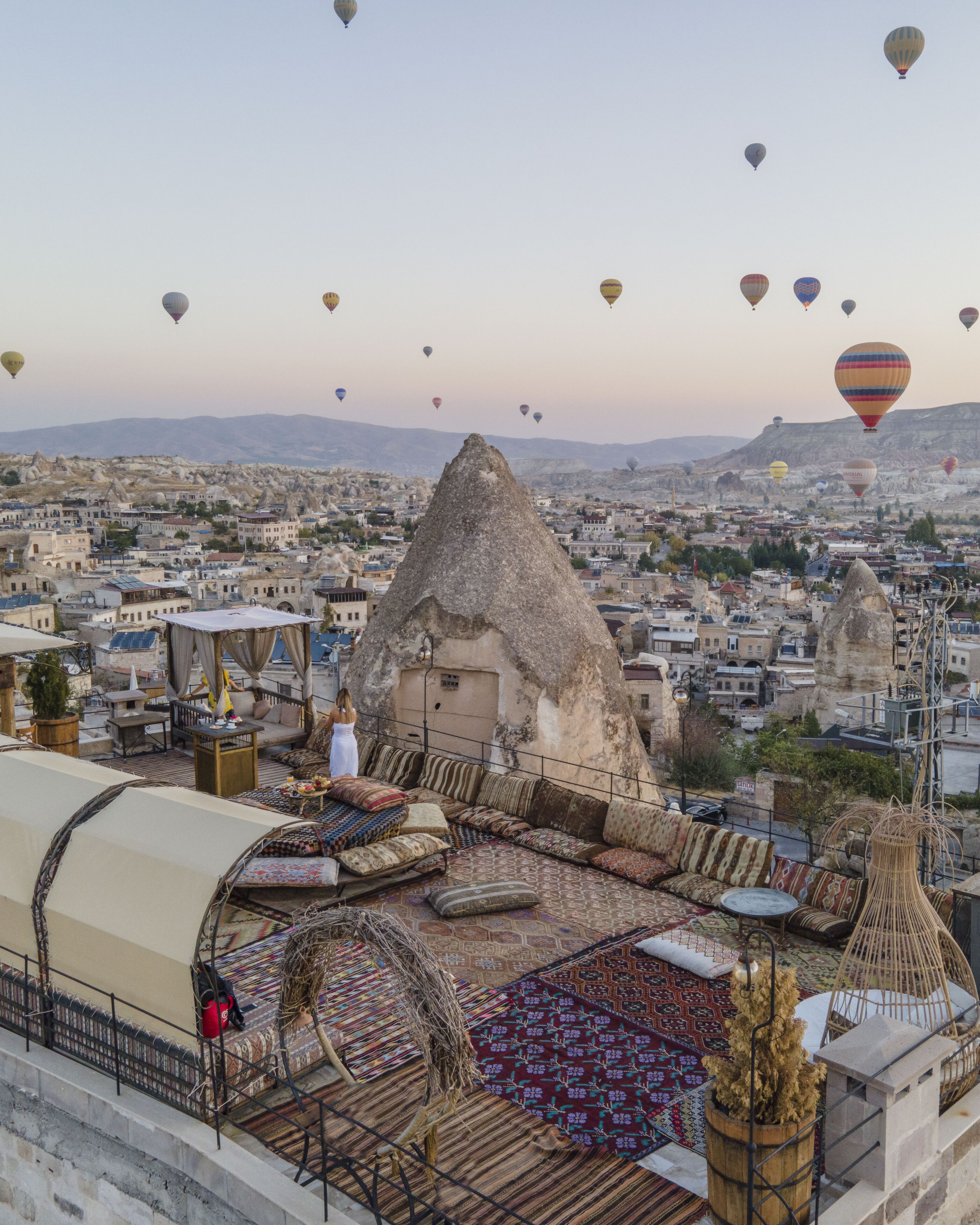 Eine Dachterrasse mit einer strohgedeckten Konstruktion, einer Sitzecke und Blick auf die Stadt sowie Heißluftballons.