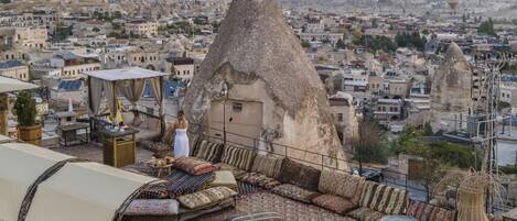A rooftop terrace with a thatched roof structure, seating area, and a view of the cityscape and hot air balloons.