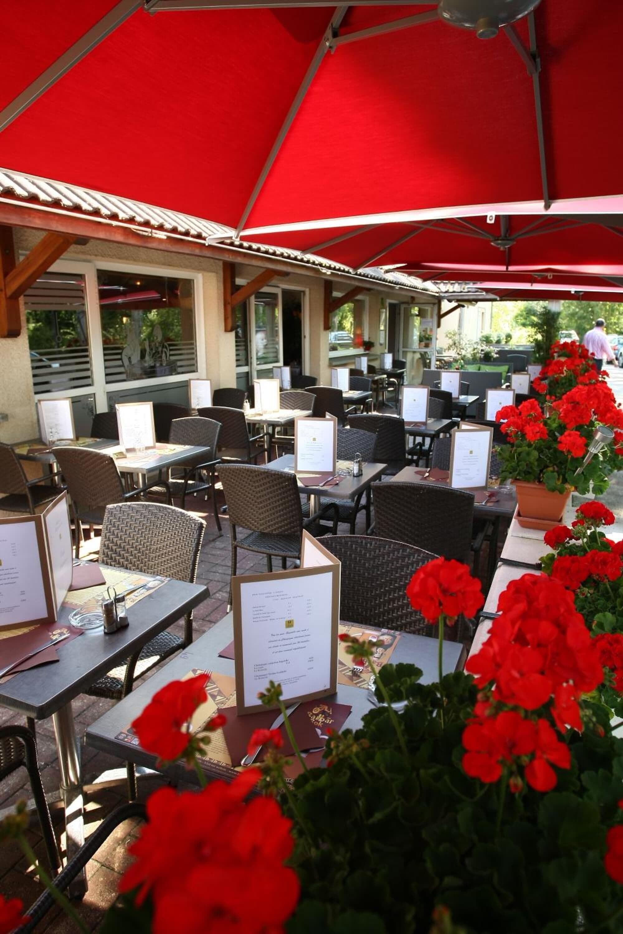 An outdoor cafe with tables and chairs under red umbrellas, surrounded by potted plants.