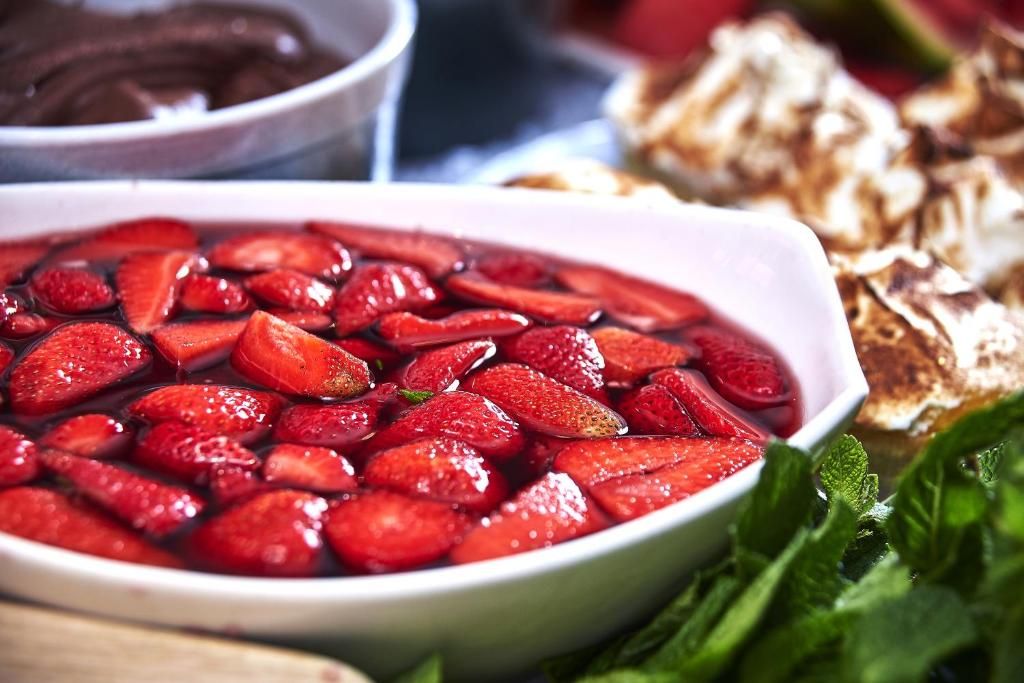 Eine Schüssel mit geschnittenen Erdbeeren, eine weiße Schüssel und ein Teller, auf dem offenbar ein Dessert steht, sind im Hintergrund zu sehen.