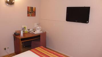 A hotel room with a bed, a desk with a telephone, a coffee maker, a painting on the wall, and a flat-screen TV.