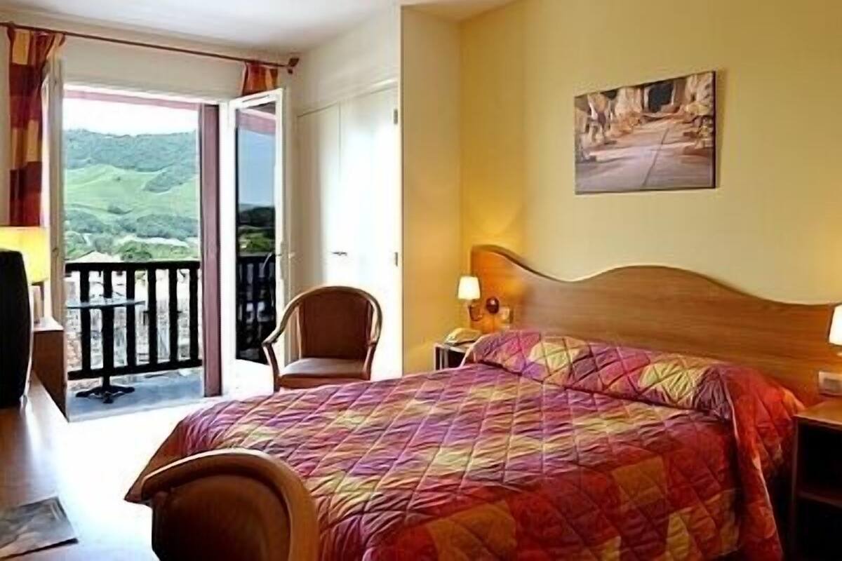 A hotel room with a bed, a chair, a nightstand, a painting on the wall, and a balcony with a view of the hills.