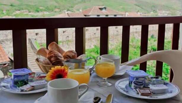 Una mesa de desayuno con cruasanes, una cafetera, un vaso de jugo de naranja y una flor en un balcón con vistas a un paisaje montañoso.