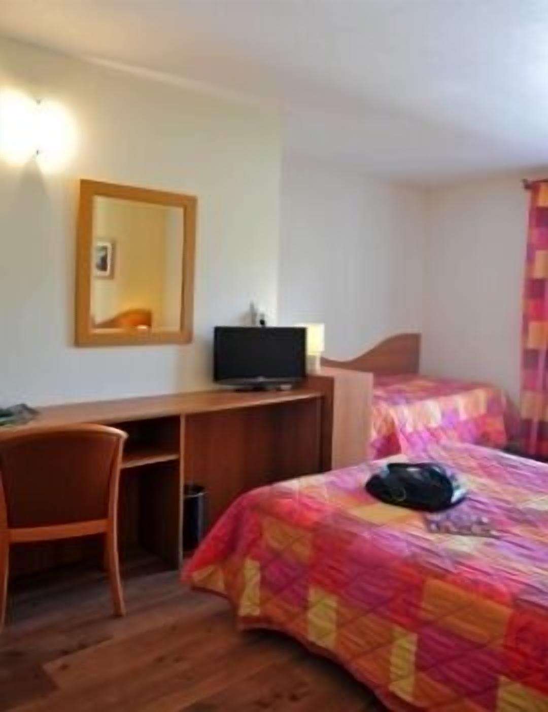 Une chambre d’hôtel avec un bureau en bois, une télévision, un lit recouvert d’une couverture aux couleurs vives et une chaise.