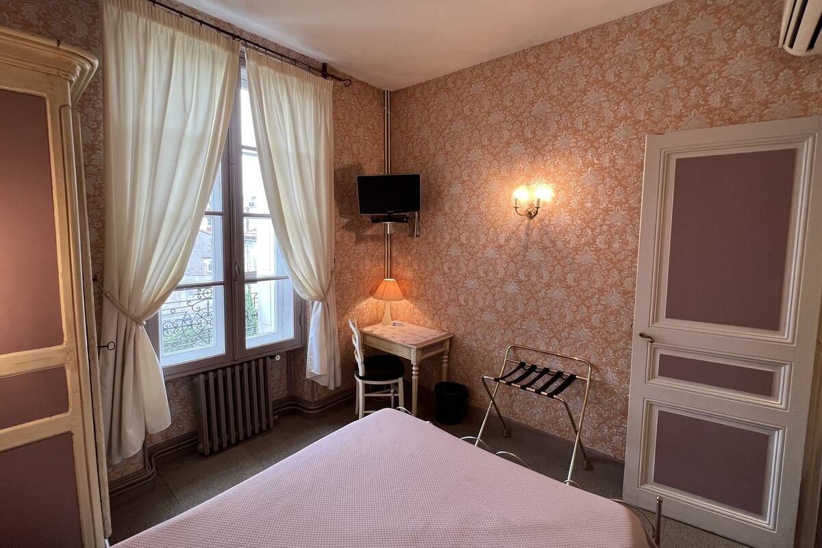 A hotel room with a bed, a desk with a lamp, a TV, and a radiator.