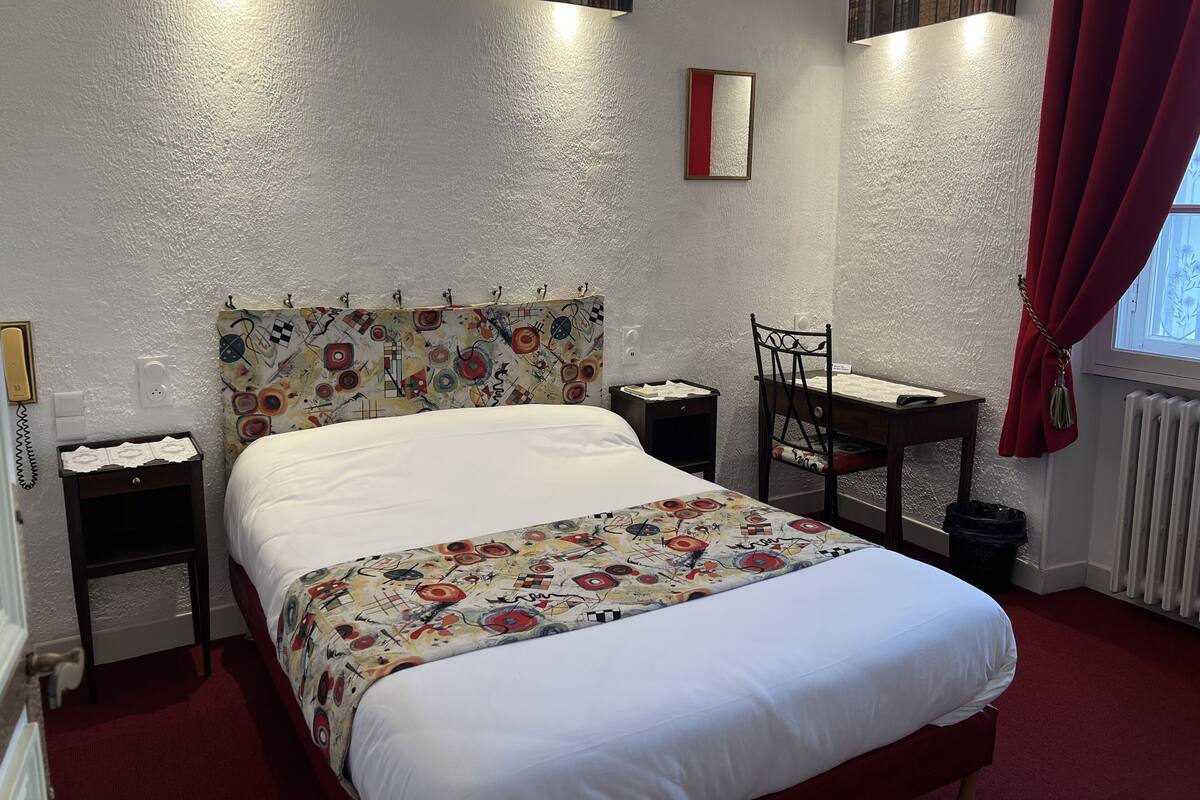A hotel room with a bed, a desk, a chair, and a window with red curtains.