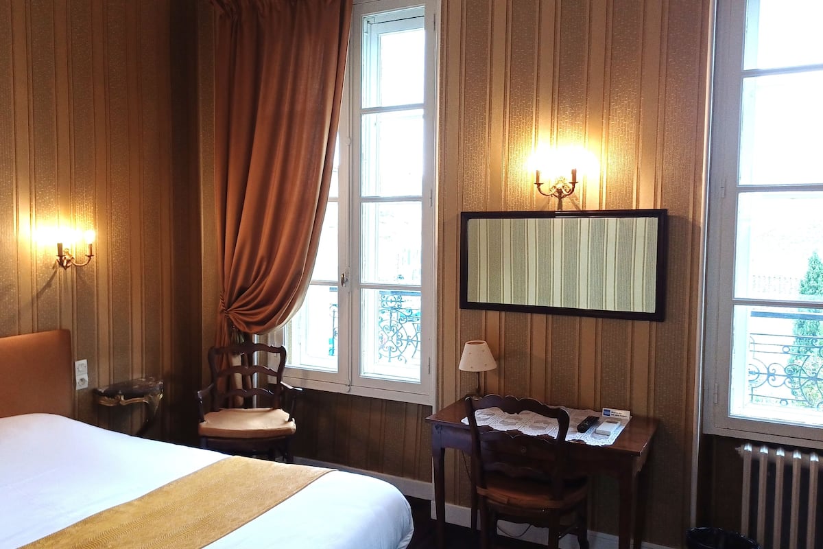A hotel room with a bed, a desk, a chair, a lamp, a wall-mounted mirror, and a window with curtains.