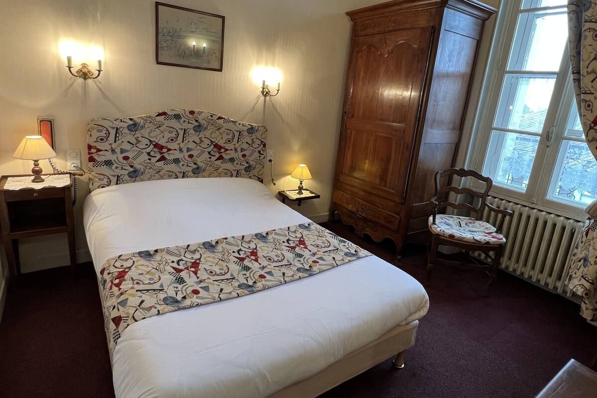 A bedroom with a bed, two bedside lamps, a wooden wardrobe, a chair, and a window with curtains.