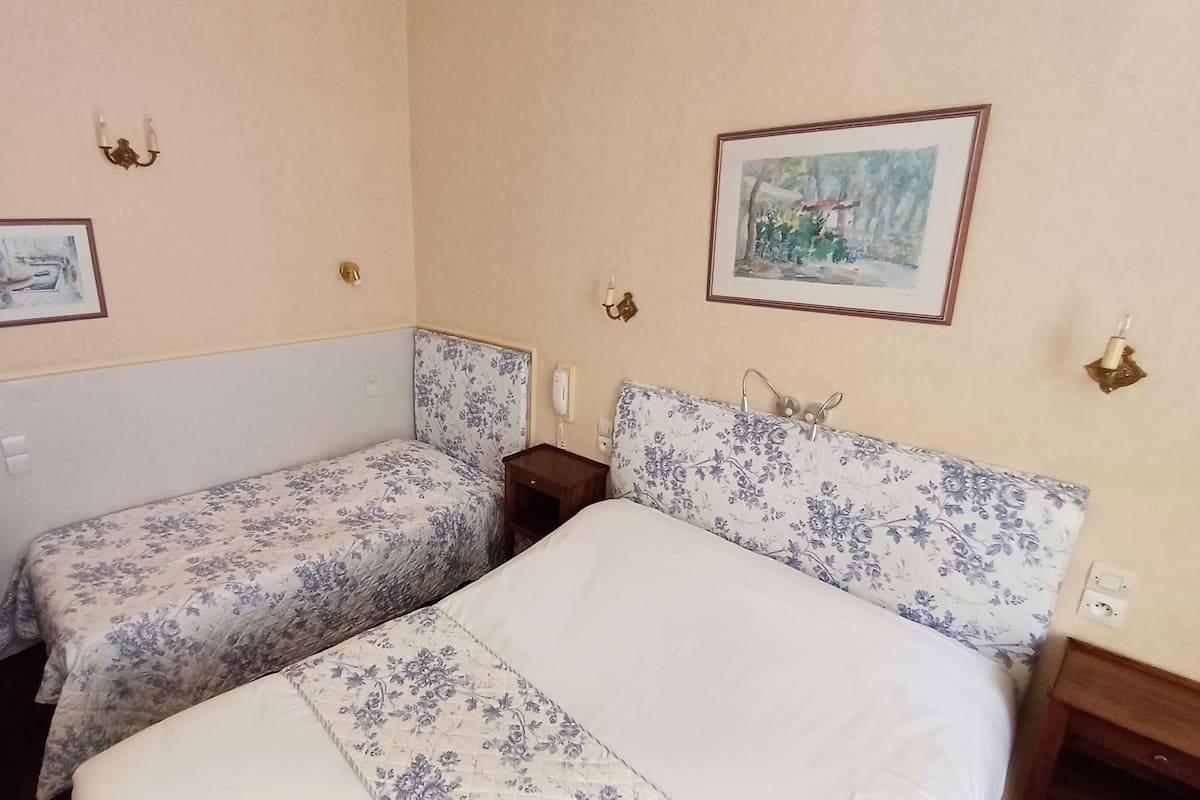 A room with two beds, one with a floral headboard and the other with a plain headboard. There are two wall-mounted light fixtures and a framed painting above the beds.