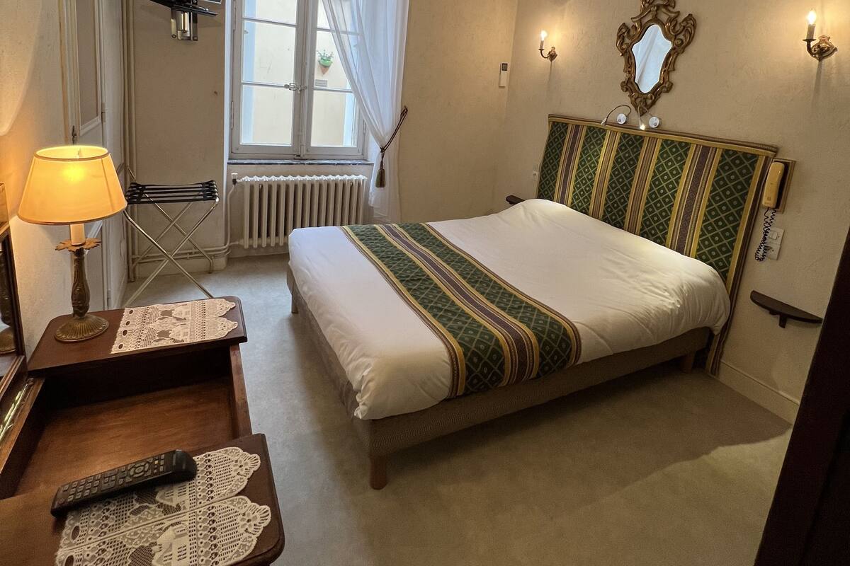 A hotel room with a bed, a desk with a lamp, a TV mounted on the wall, and a window with curtains.