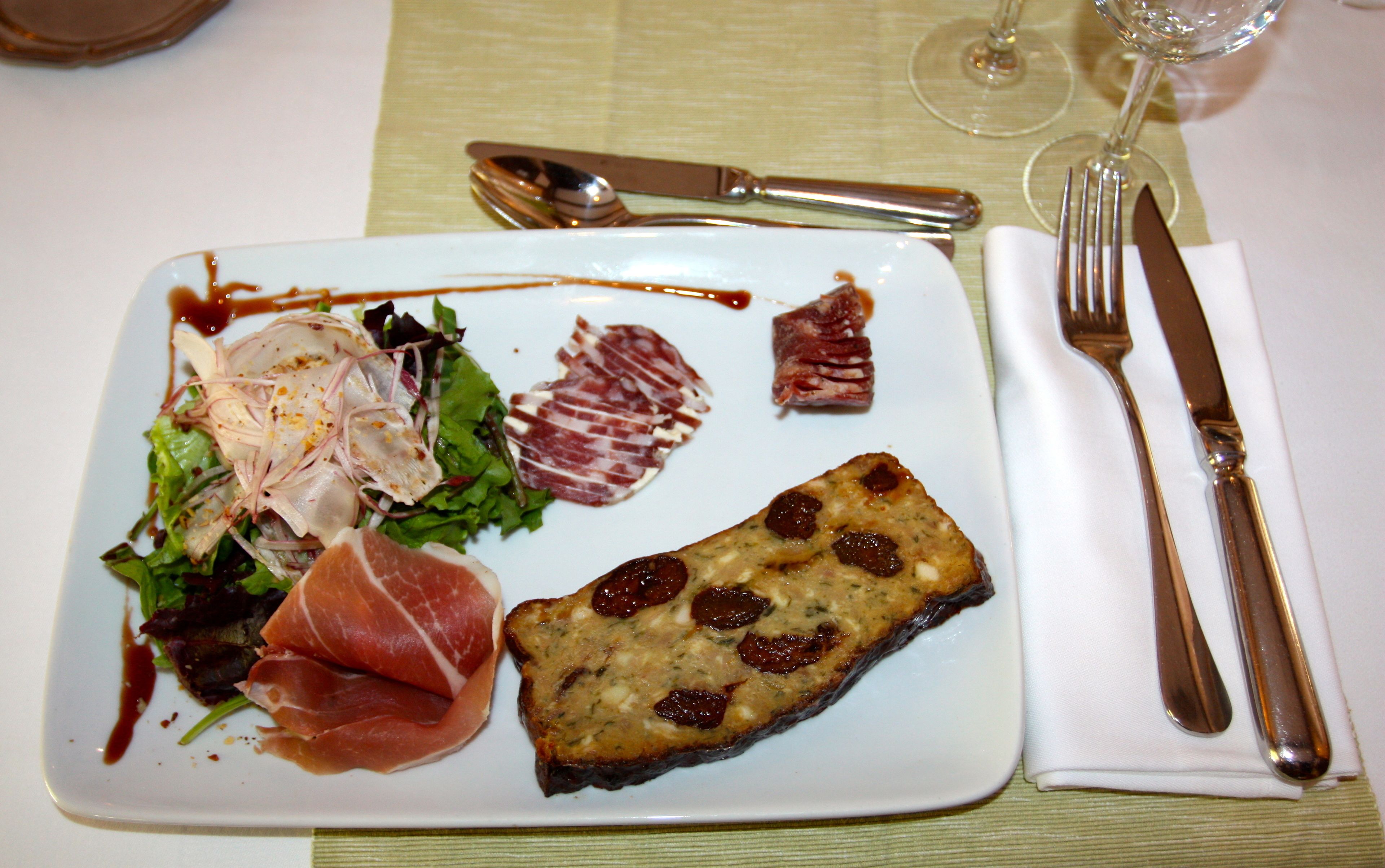 A plate of food with various meats and a slice of bread, accompanied by a fork and knife on a tablecloth.