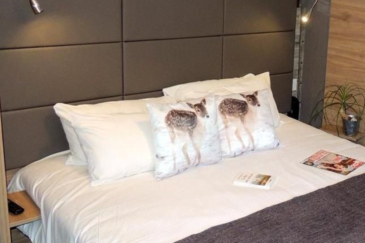 A neatly made bed with white bedding and a brown throw blanket, a bedside table with a lamp, and a wall-mounted coat rack.