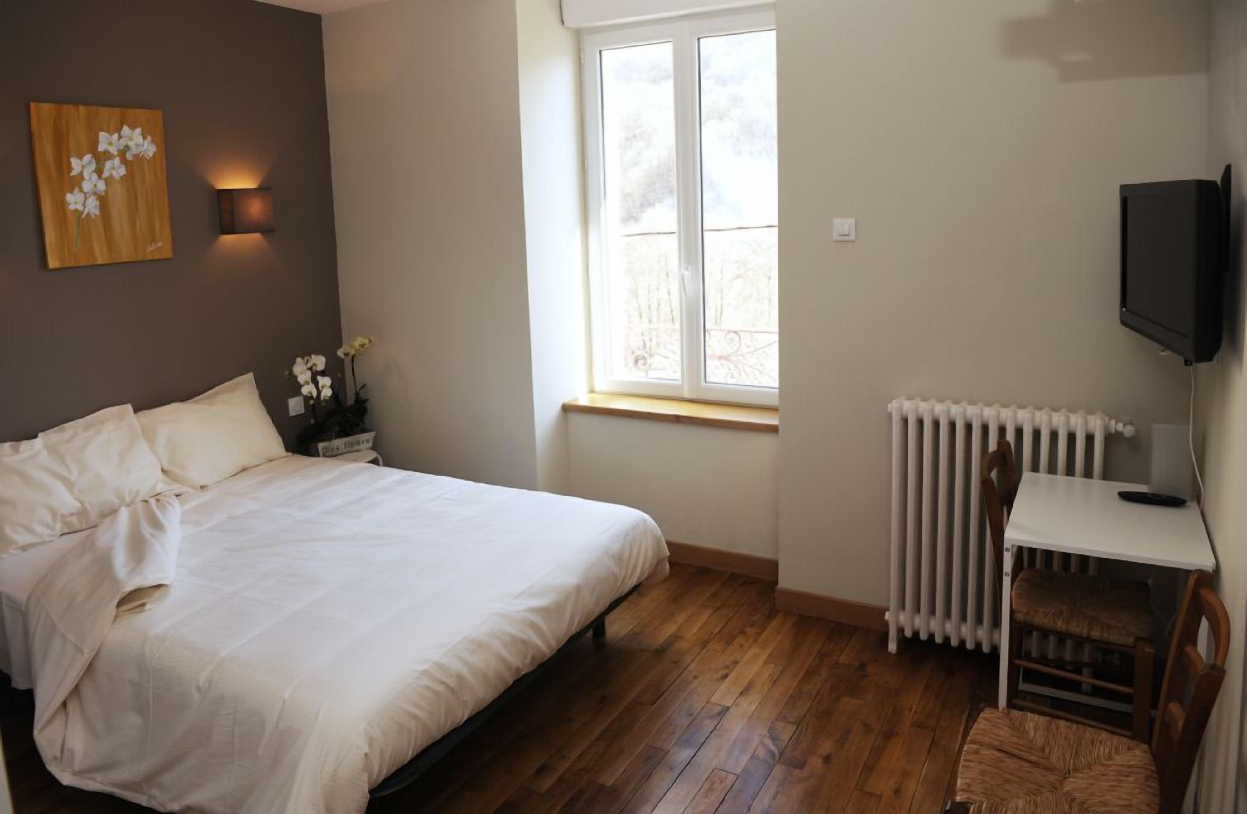 Ein Schlafzimmer mit Bett, kleinem Tisch, Stuhl, Heizkörper, Fernseher und Fenster mit Blick auf Bäume.