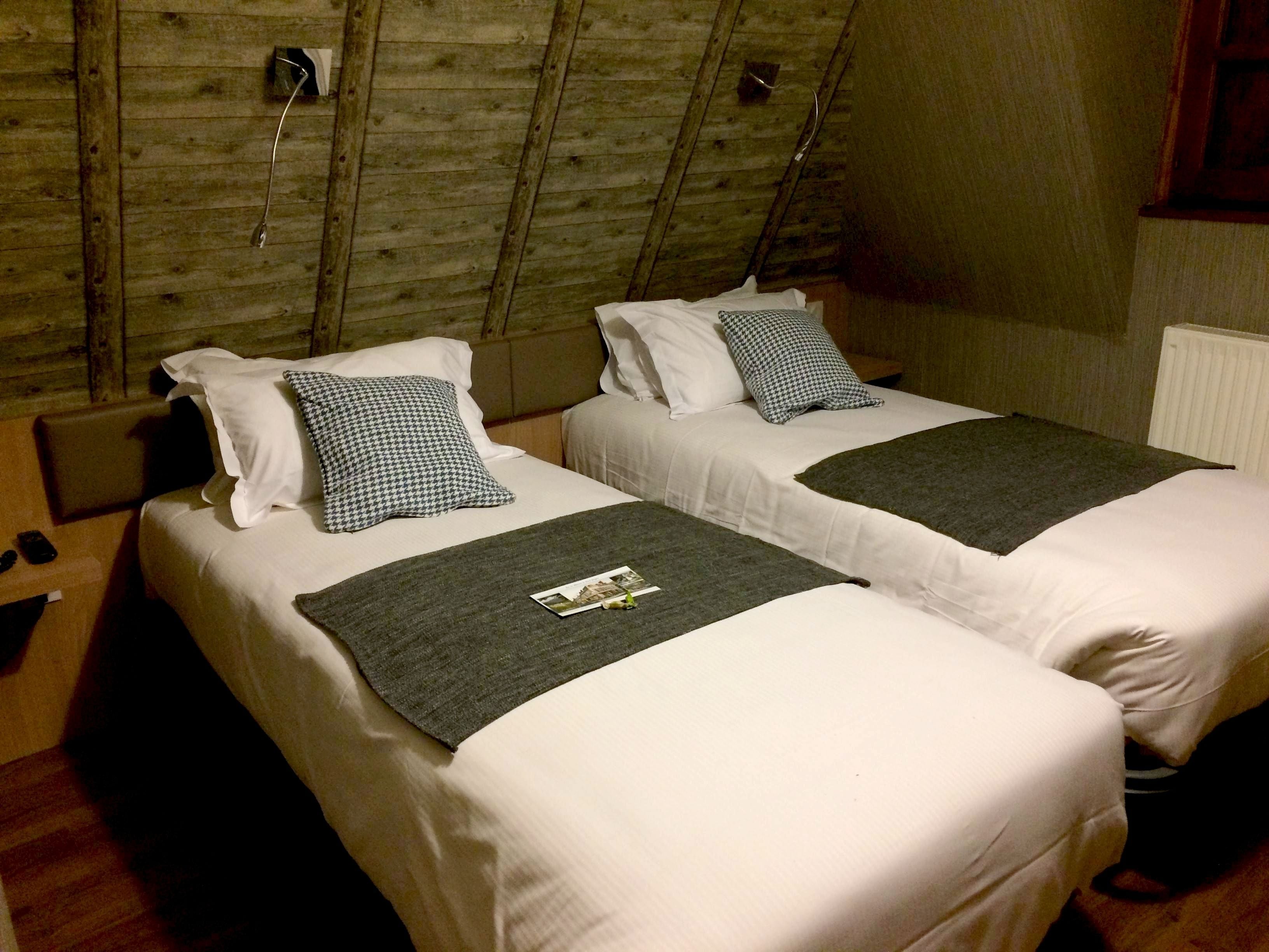 Deux lits simples avec du linge de lit blanc et des coussins à motif pied-de-poule, un plaid gris foncé et une petite table avec une télécommande.