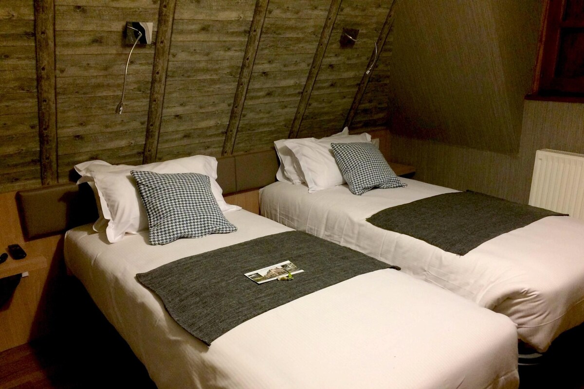 Dos camas con sábanas blancas y almohadones de piedritas, una manta de lana gris y techo de madera.