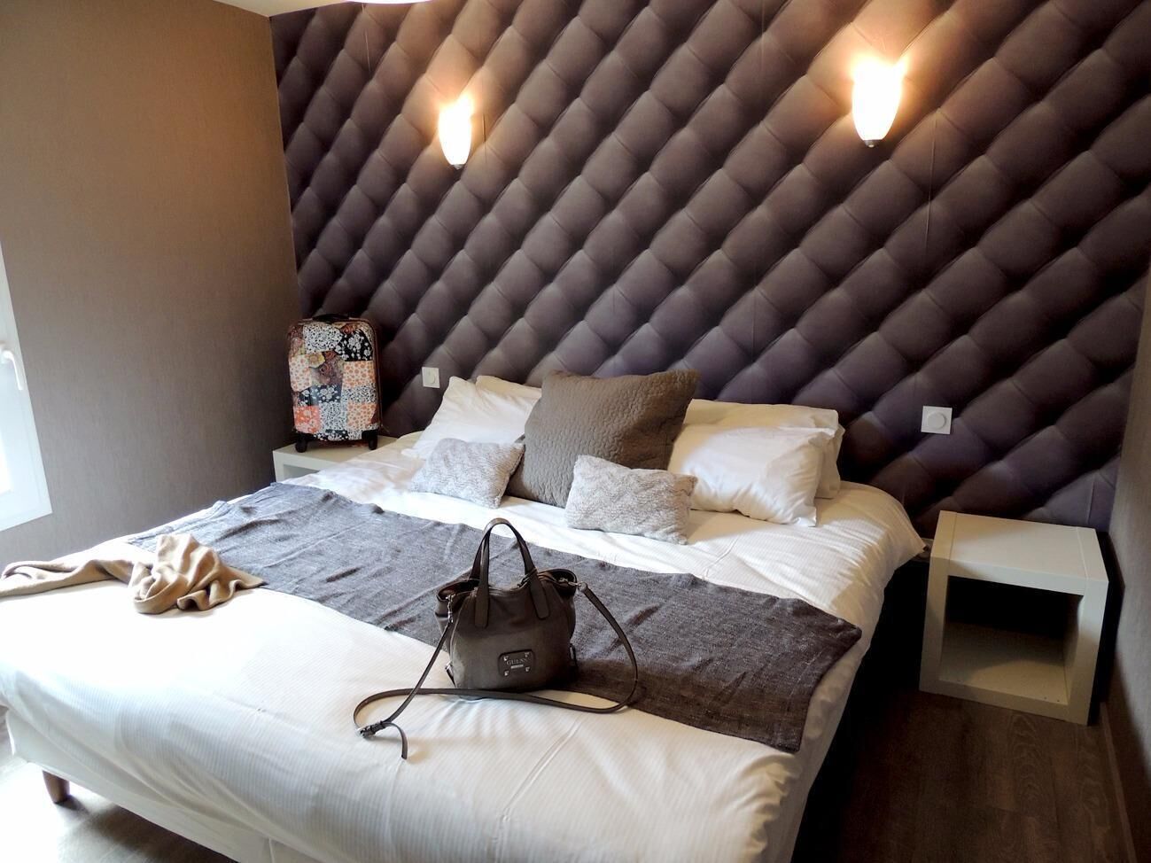 Un dormitorio moderno con cabecera capitonada, cama con sábanas blancas, manta gris, bolso marrón y dos mesitas de noche.