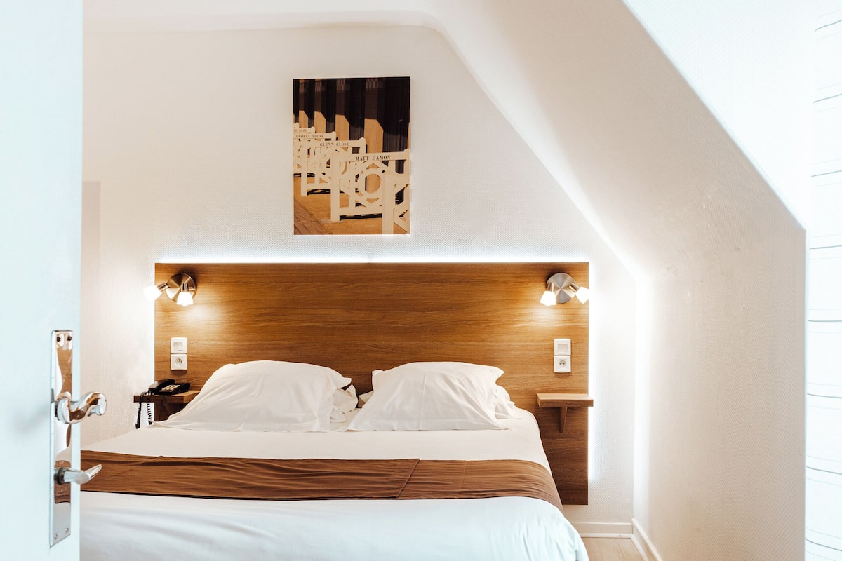 A bedroom with a wooden headboard, a bed with white bedding, two bedside tables, and a wall-mounted light fixture.
