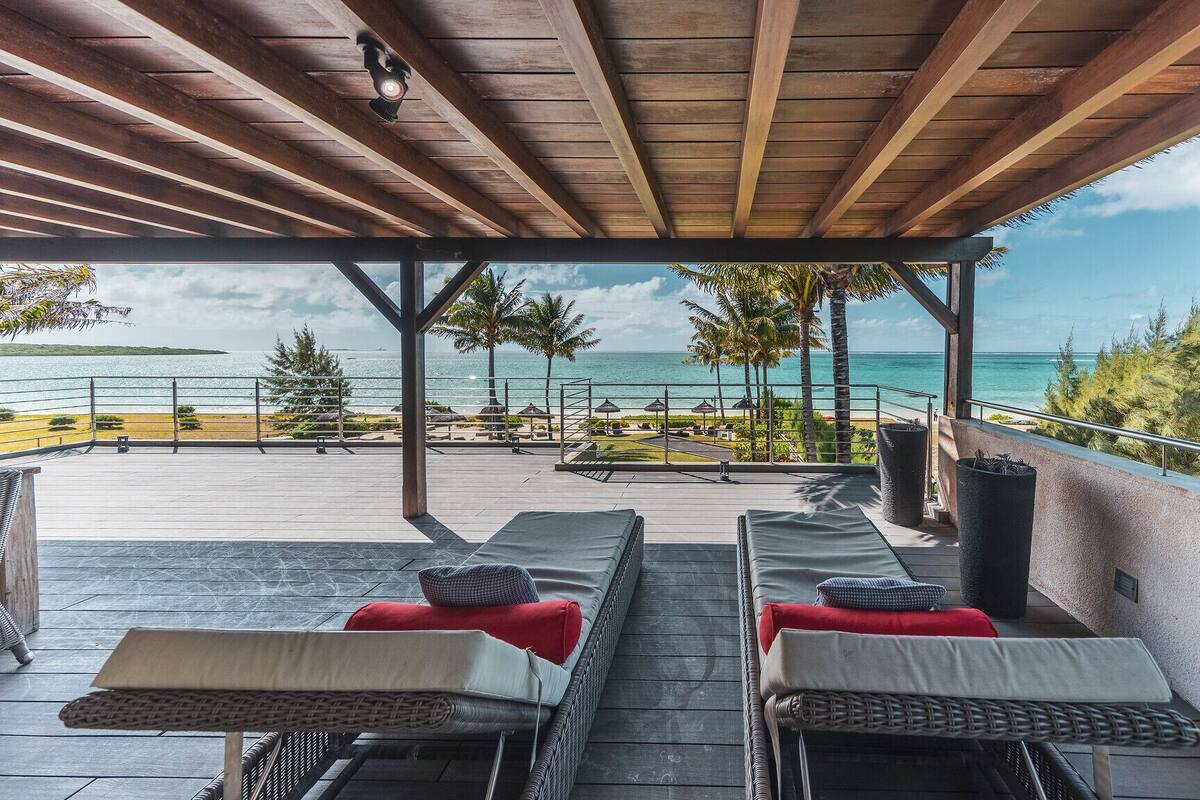 Un espace extérieur aménagé avec des meubles de jardin en osier et des coussins rouges, offrant une vue sur une plage et l’océan.