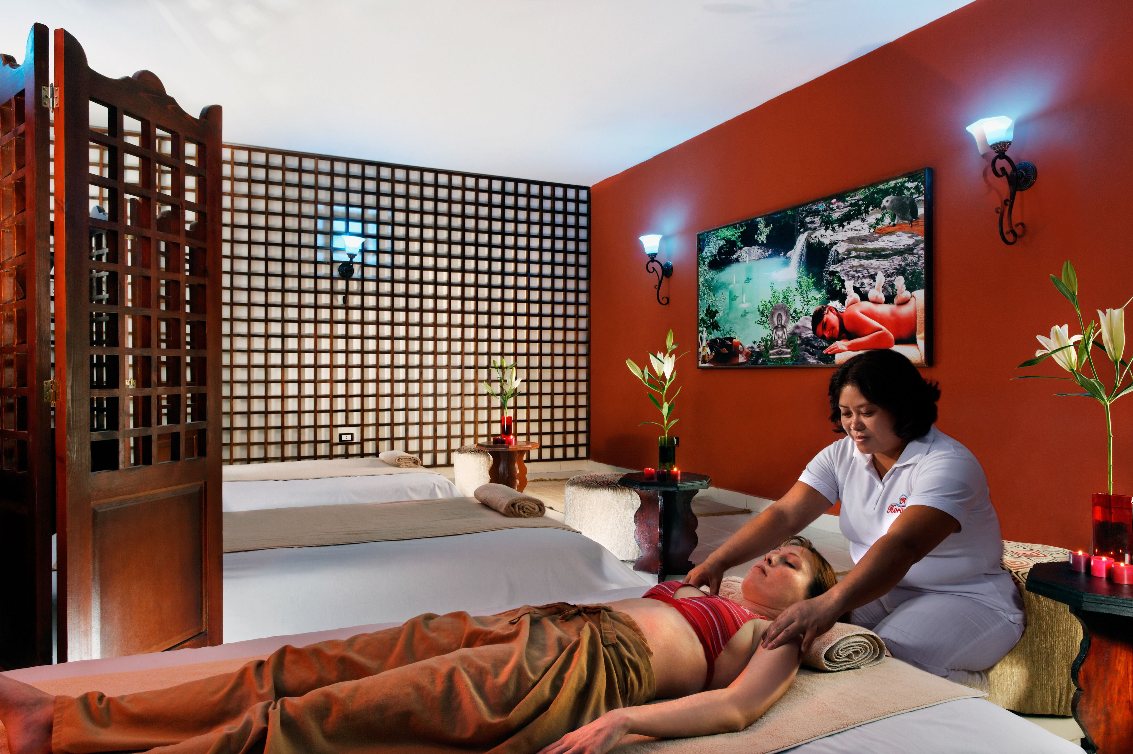 Een massageruimte met een houten scheidingswand, een bed, een massagetafel, een schilderij aan de muur en twee wandlampen.