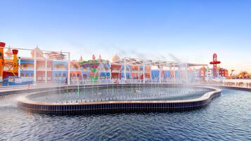 Ein Wasserbrunnen mit kreisförmigem Becken, umgeben von einem weißen Zaun und farbenfrohen Gebäuden im Hintergrund.