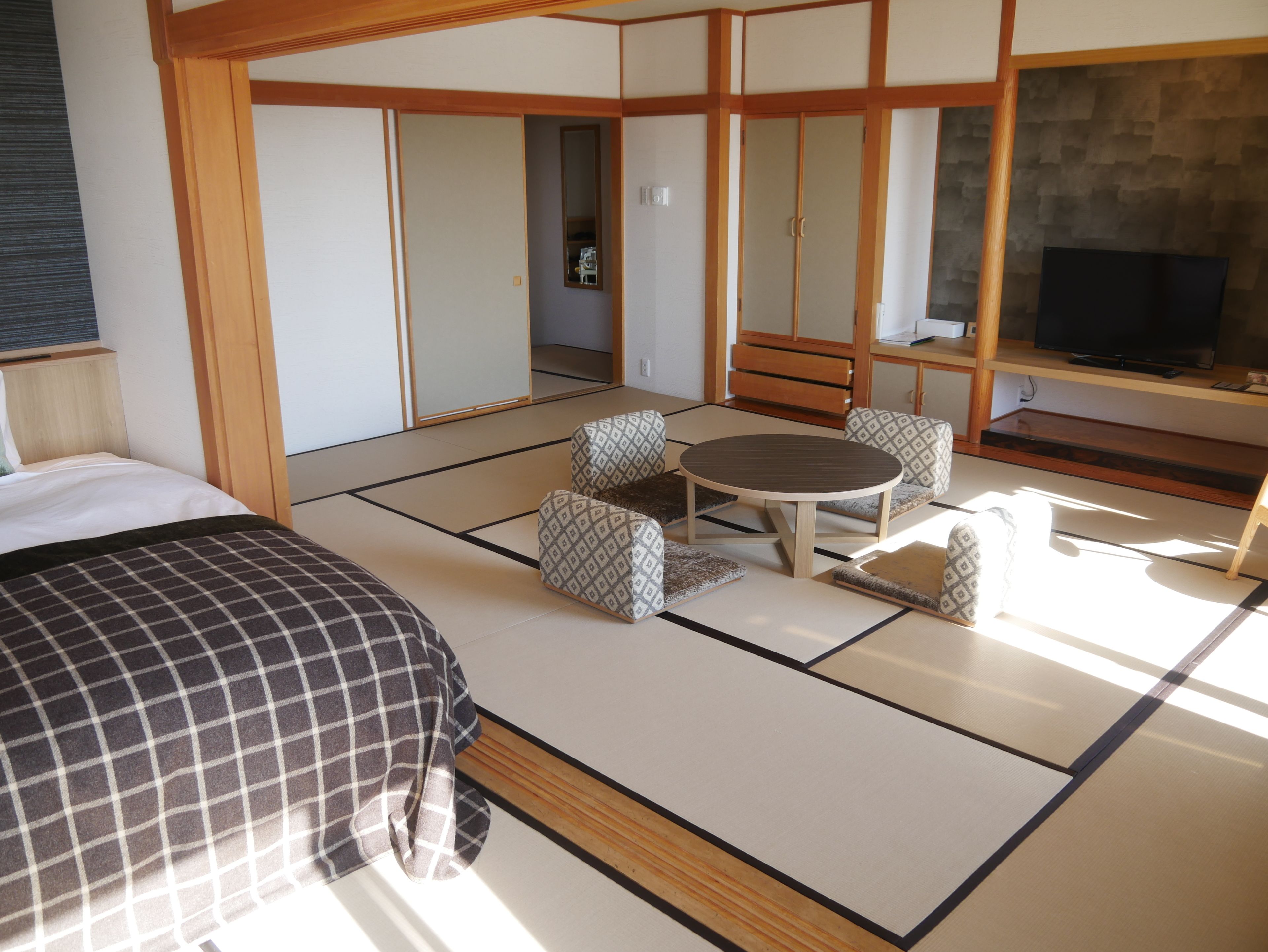 A traditional Japanese-style room with tatami flooring, a low table with cushions, a bed with a checkered bedspread, and a television on a wooden stand.