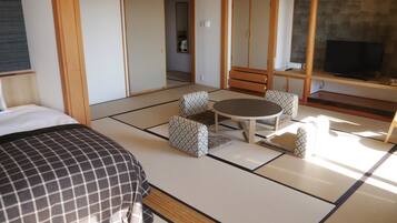 A traditional Japanese-style room with tatami flooring, a low table with cushions, a bed with a checkered bedspread, and a television on a wooden stand.