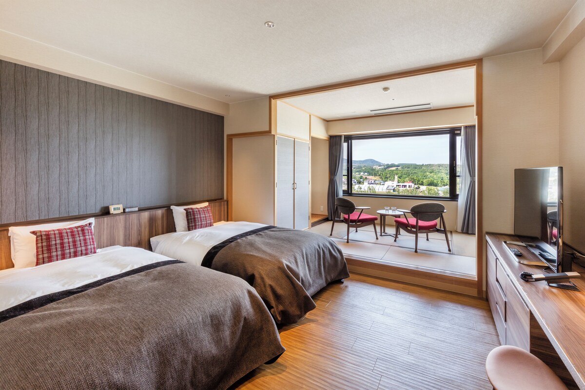 A hotel room with two beds, a desk, a television, and a view of a cityscape.