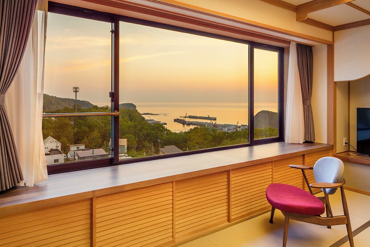 A room with a large window overlooking a body of water and a distant landmass, a wooden bench, and a television.