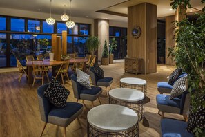 A modern lounge area with wooden flooring, blue upholstered chairs, round tables, and large windows offering a view of the cityscape at dusk. - Kiki Shiretoko Natural Resort (Shari)
