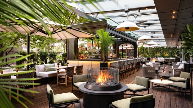 An outdoor seating area with a central fire feature, surrounded by cushioned chairs and tables, under a covered patio with a bar in the background. - The Ritz-Carlton, Toronto (Toronto)