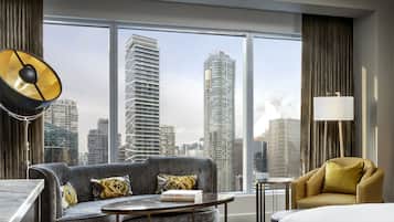 A modern hotel room with a large window offering a city view, a sofa with patterned cushions, a small table, and a lamp.