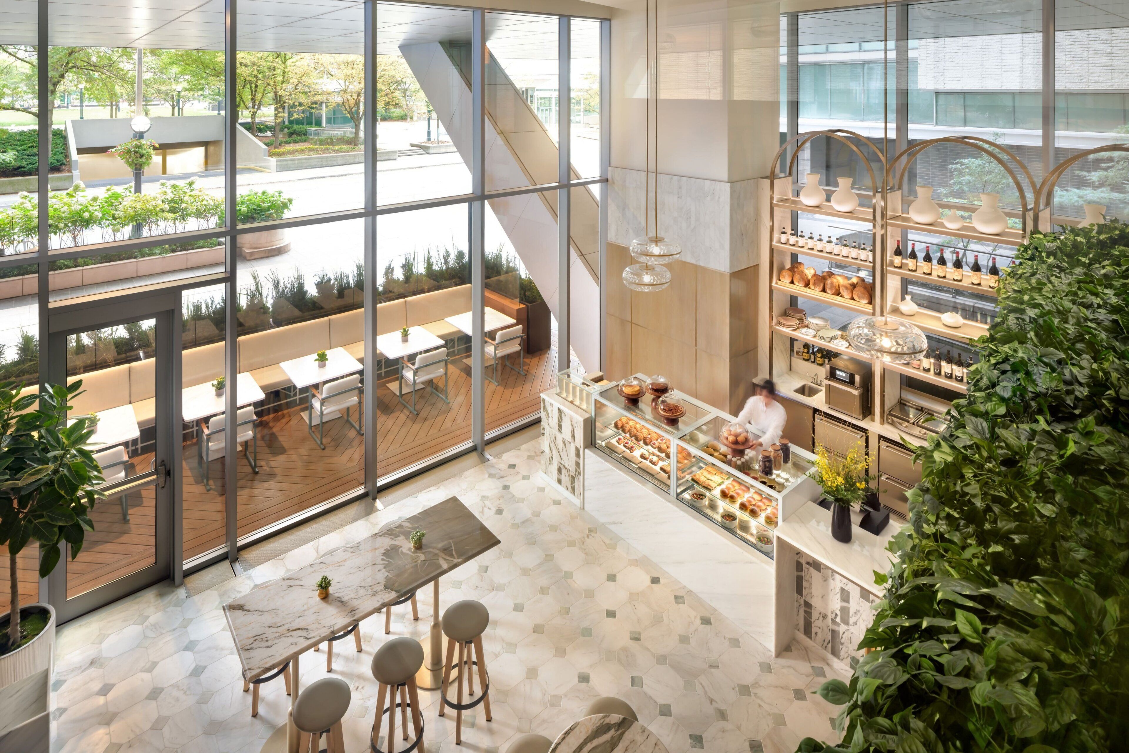 Una cafetería moderna con amplios ventanales, un espacio de estar con mesas y sillas, y una exhibición de productos horneados y bebidas.