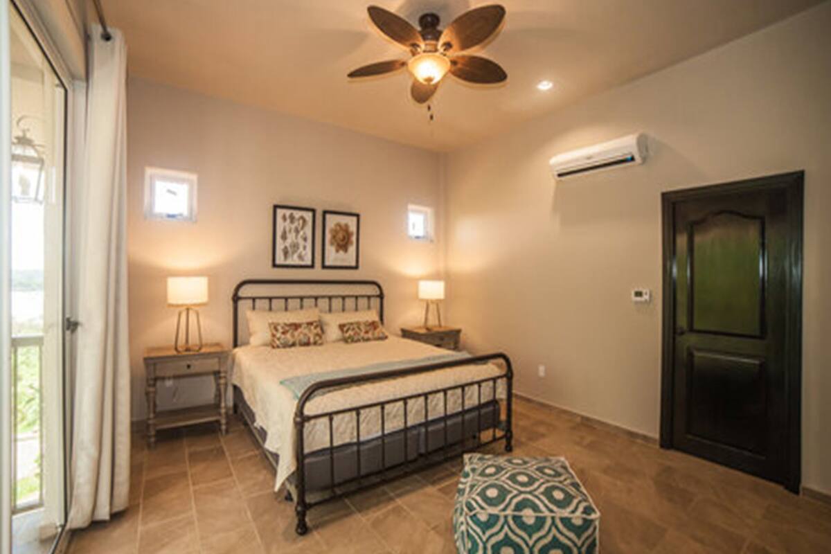 Une chambre à coucher comprenant un lit, des lampes de chevet, un ventilateur de plafond, un appareil de climatisation et une porte.