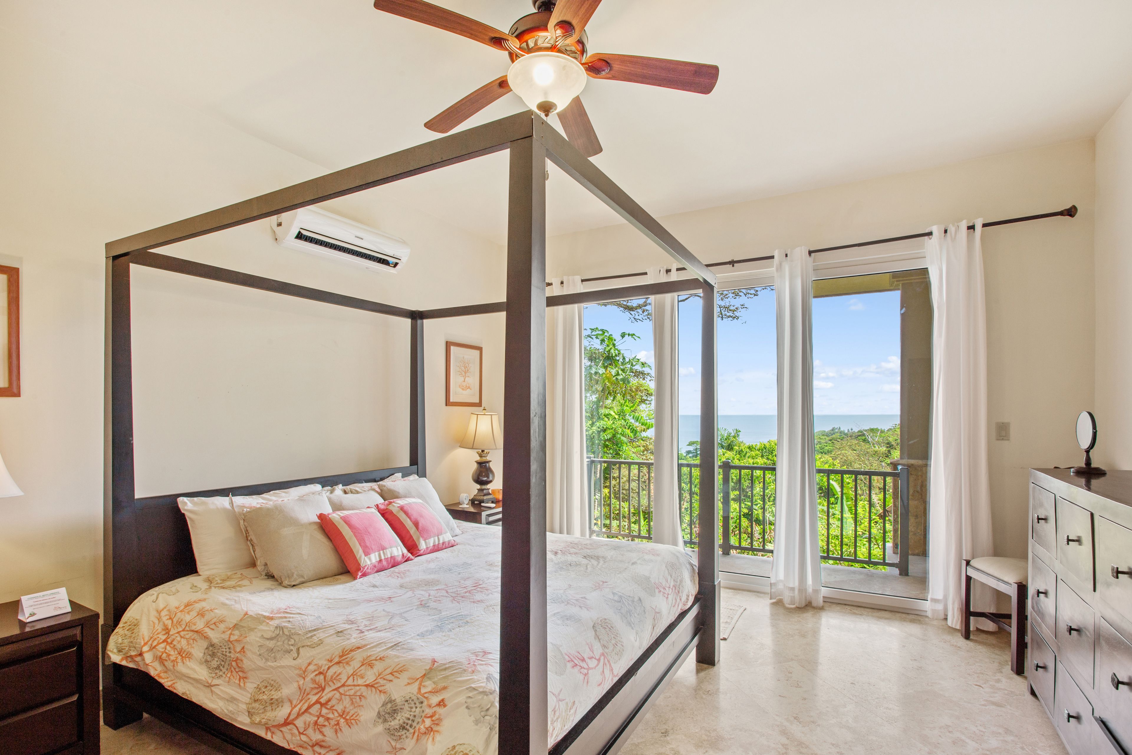 A four-poster bed with a view of the ocean, a balcony with a railing, and a dresser with a mirror.