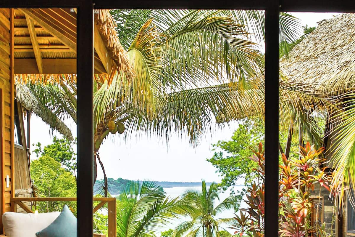 Une terrasse en bois avec une piscine à fond de verre, un canapé blanc avec un coussin bleu et une vue sur l’océan et les palmiers.
