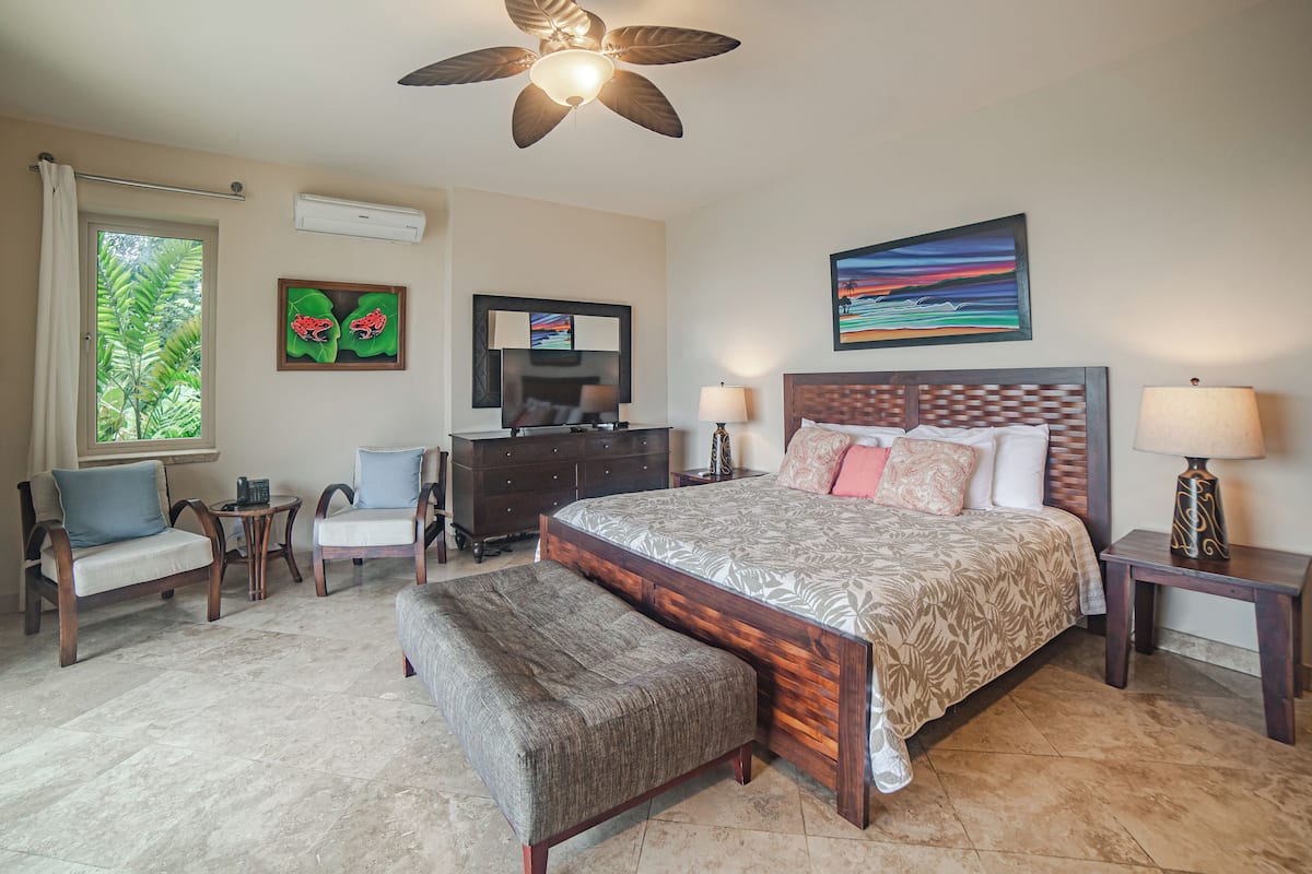 Une chambre avec un grand lit, un ventilateur de plafond, une commode, un bureau, deux chaises, un banc et une fenêtre donnant sur un paysage verdoyant.