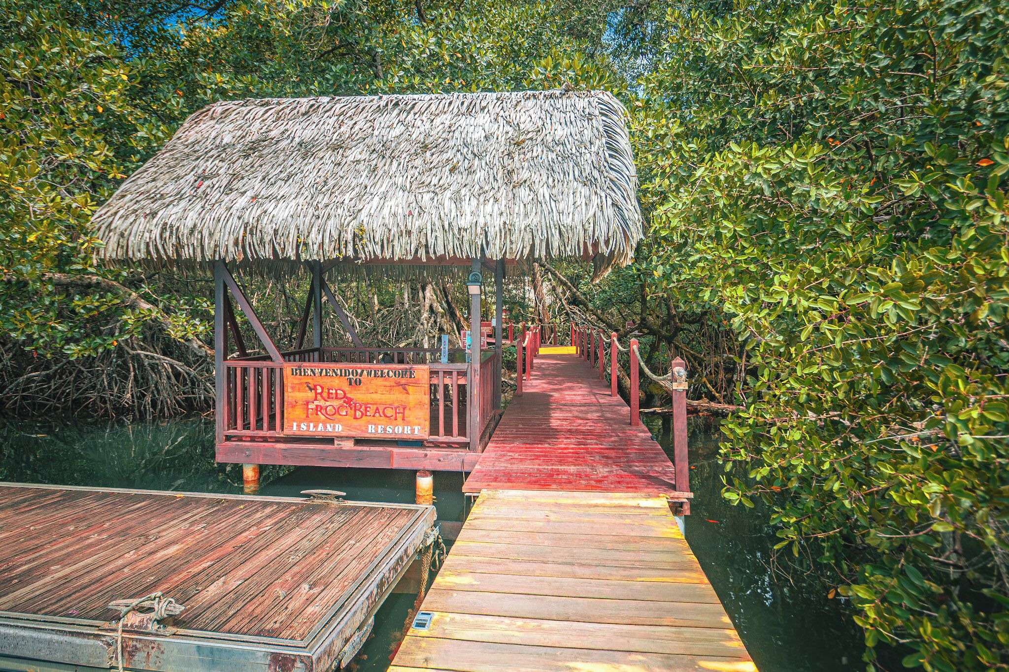 Een houten steiger die leidt naar een gebouw met een rieten dak, met een bord waarop staat: Welcome to Red Beach Island Resort.
