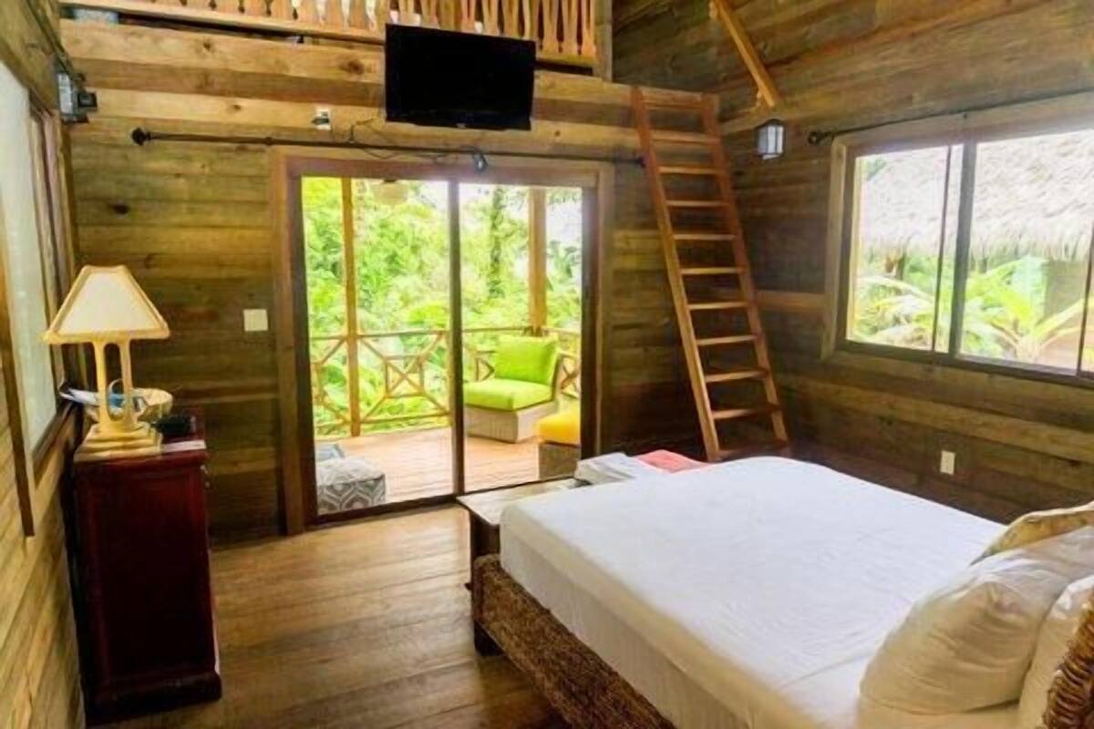 Une chambre aménagée dans une cabane en bois, avec un lit, une télévision, un bureau avec une lampe, une échelle et un balcon avec une chaise.