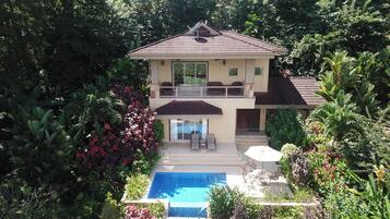 A two-story house with a swimming pool, surrounded by lush greenery.
