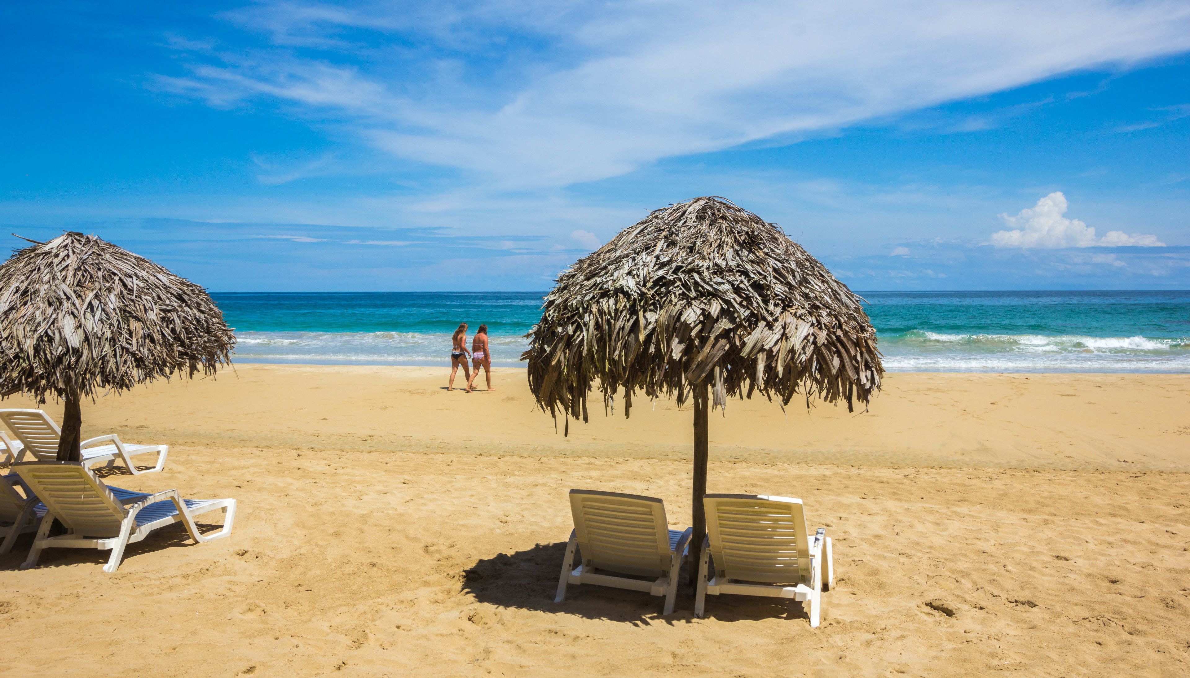 Una playa con dos sombrillas de paja y sillones blancos.