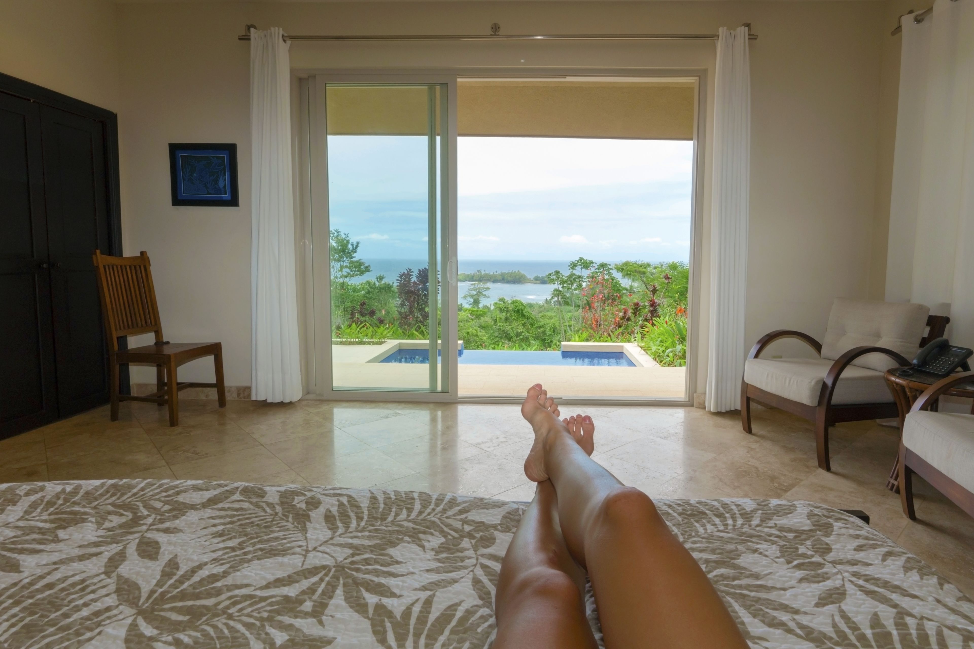 A room with a bed, a chair, a desk, and a view of a pool and greenery through a sliding door.