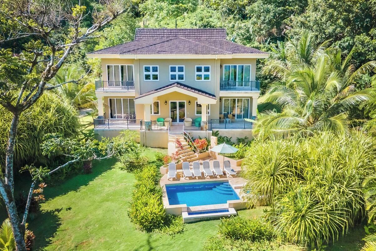 Une maison à deux étages avec une piscine, entourée d’une végétation luxuriante et de palmiers.