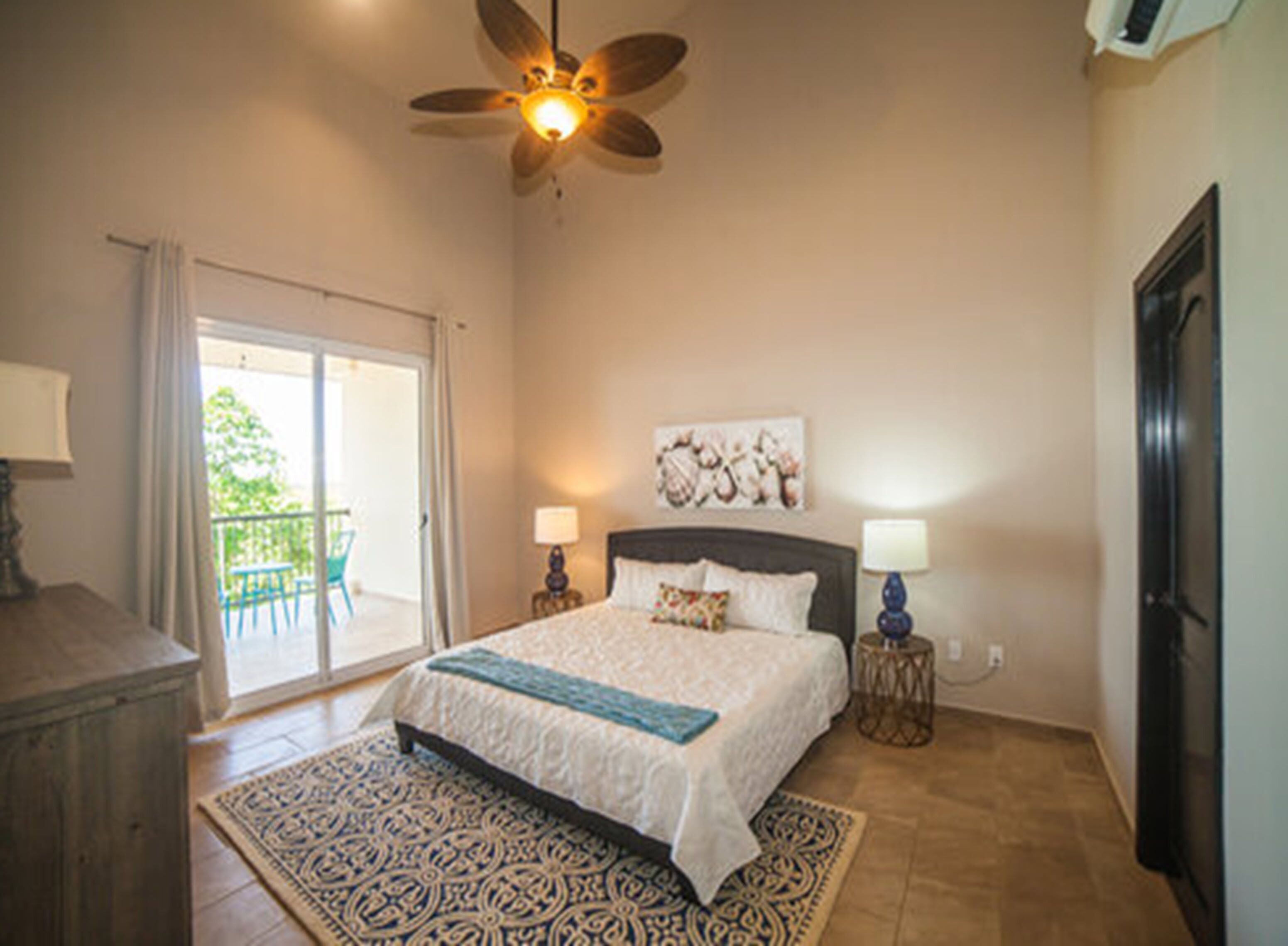 A bedroom with a bed, two bedside lamps, a ceiling fan, a large window with a view, and a door leading to a balcony.
