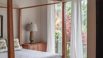 A four-poster bed with a wooden frame, a bedside table with a lamp, and a view of a balcony with red foliage.