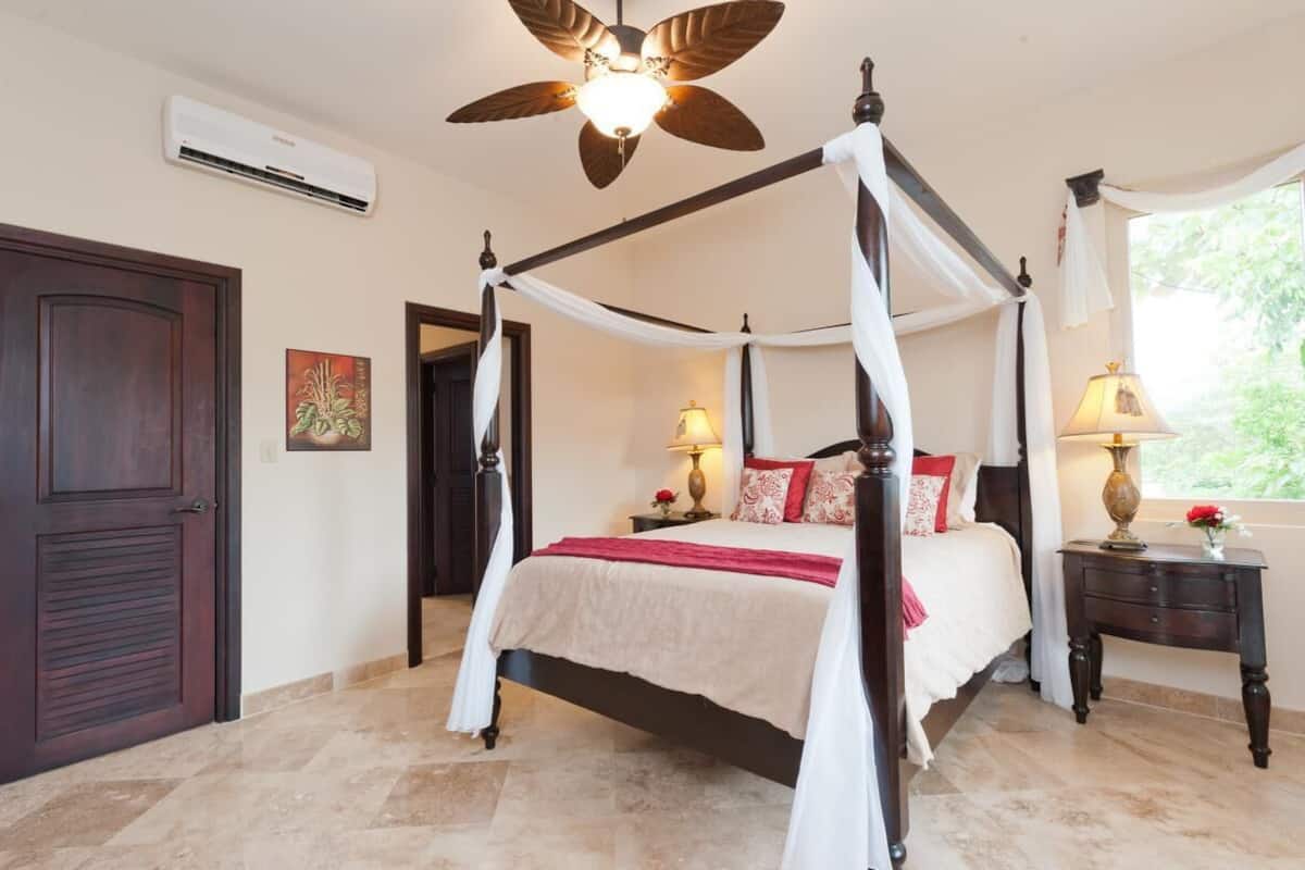 Un lit à baldaquin, un ventilateur de plafond, un appareil de climatisation, une table de chevet avec une lampe et une fenêtre donnant sur un paysage verdoyant.