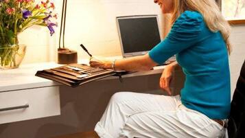 Une personne assise à un bureau, devant un ordinateur portable, avec une lampe et un vase de fleurs.