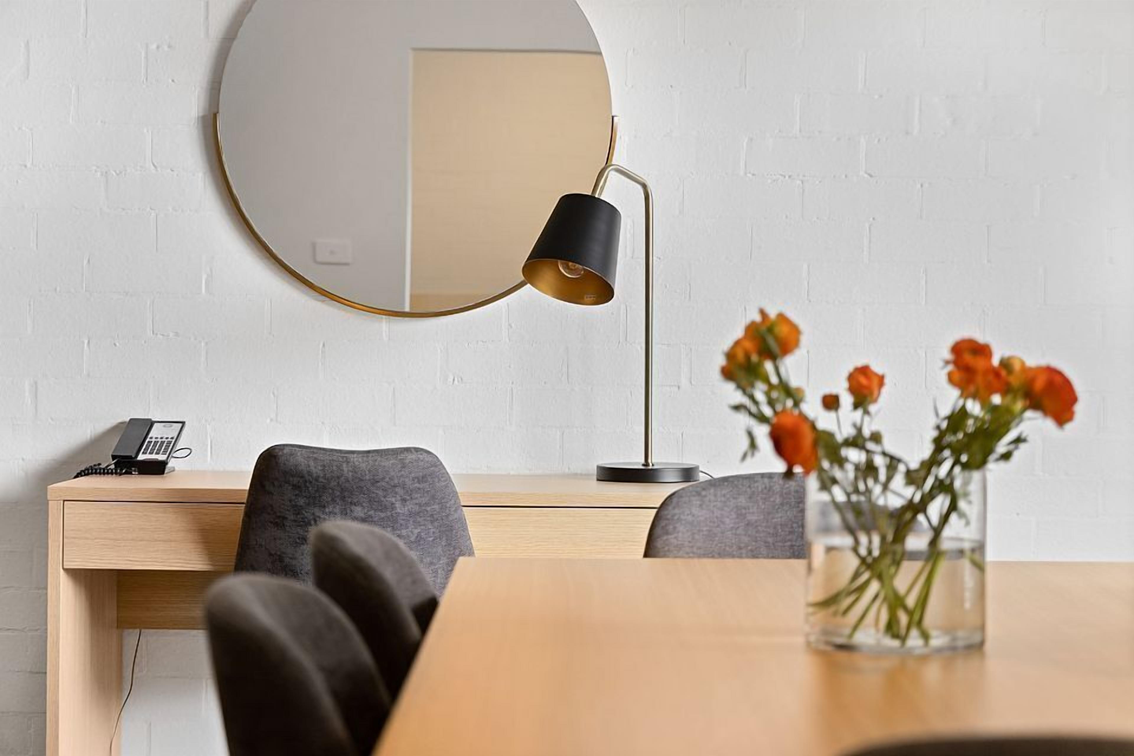 A modern office space with a round mirror, a desk with a telephone, two chairs, and a vase with flowers.