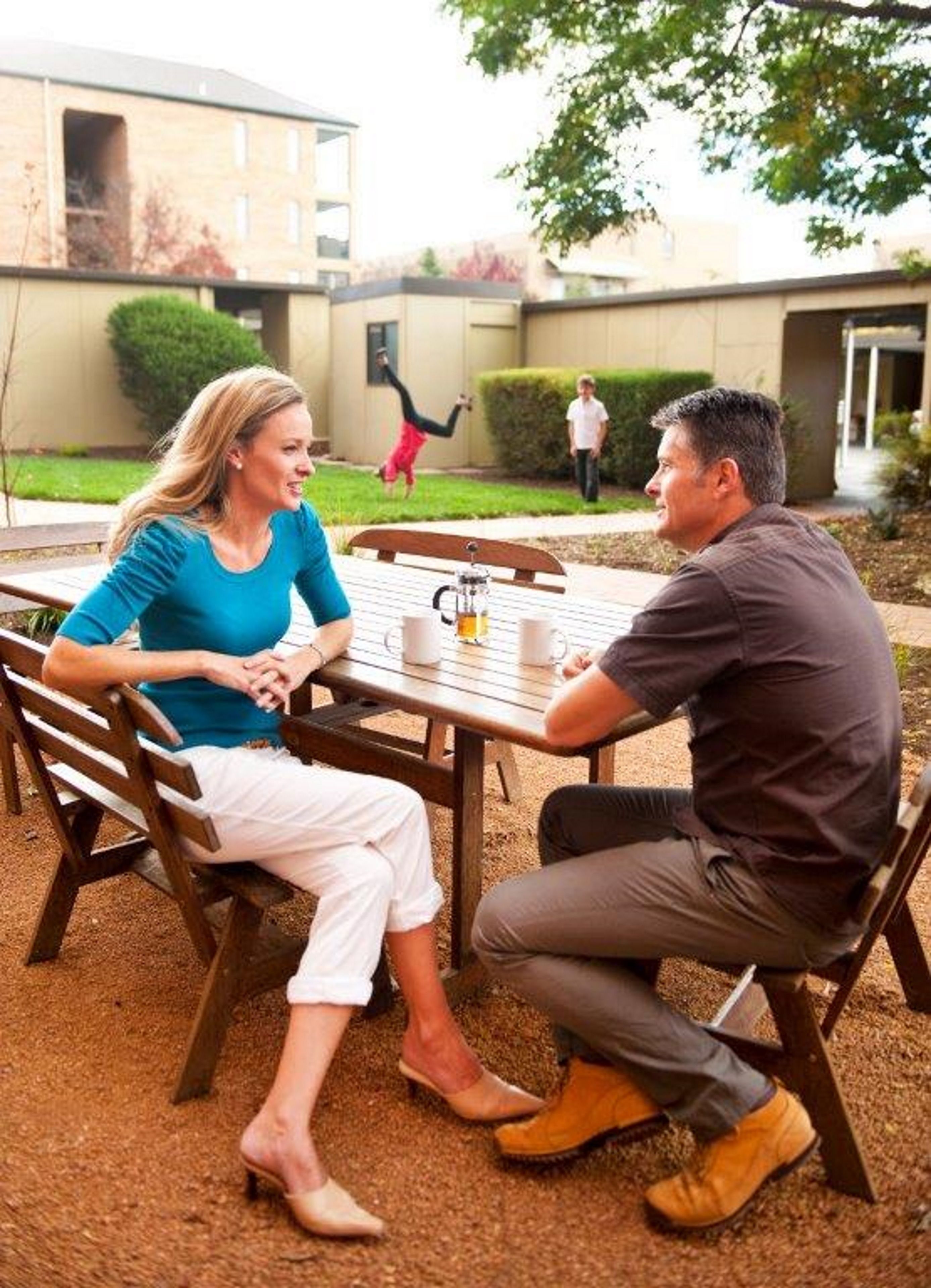 Zwei Personen sitzen an einem Tisch im Freien mit einer Kaffeekanne, in einem Innenhof mit einem Gebäude im Hintergrund.